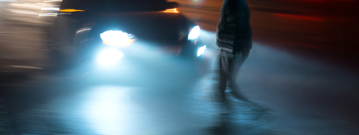 Image shows a young man crossing the road on a dark day and a speeding car approaches him