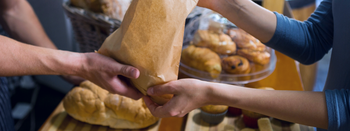 A woman hands a brown paper bag to a man. There are pastries in the background.