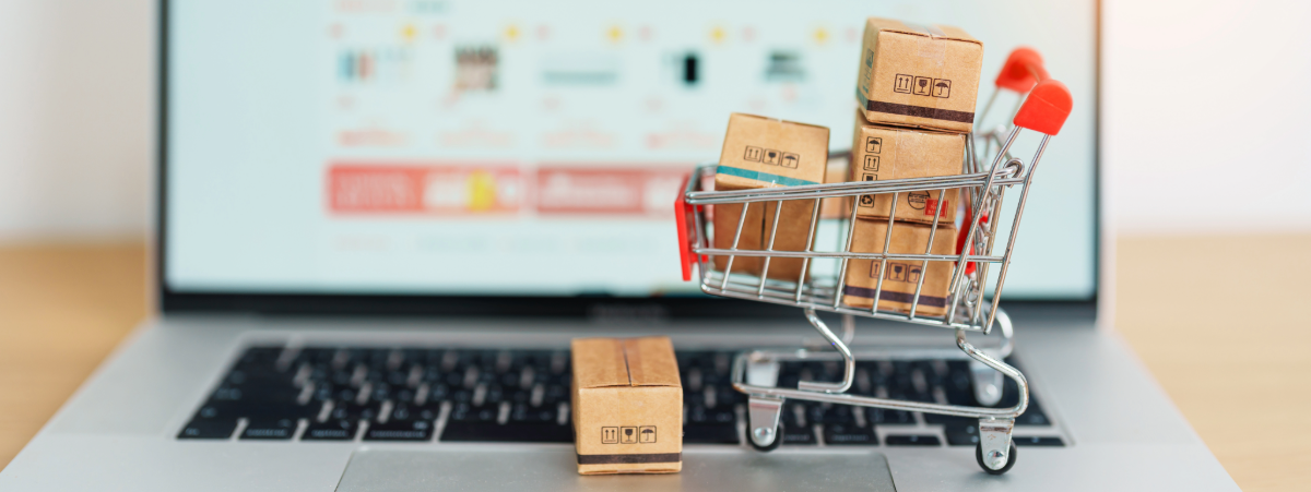 A laptop pictured on an online shopping page - blurred. A small shopping trolley sits on the keyboard filled with small boxes.