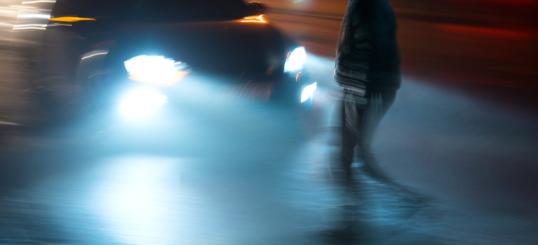 Image shows a young man crossing the road on a dark day and a speeding car approaches him