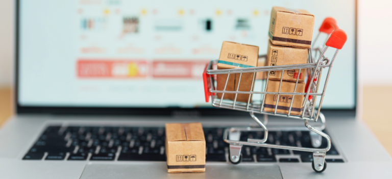 A laptop pictured on an online shopping page - blurred. A small shopping trolley sits on the keyboard filled with small boxes.